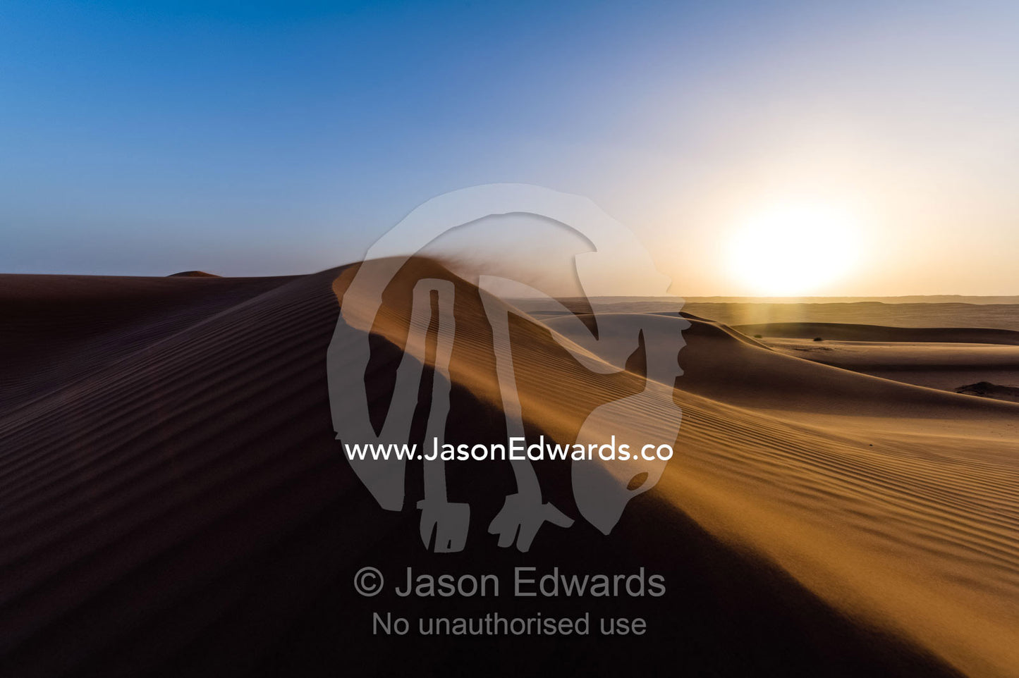 Sunset caressing the crests of red sand dune waves and howling wind in the desert. Wahiba Sands, Ramlat al-Wahiba, Sharqiya Sands, Sultanate of Oman.
