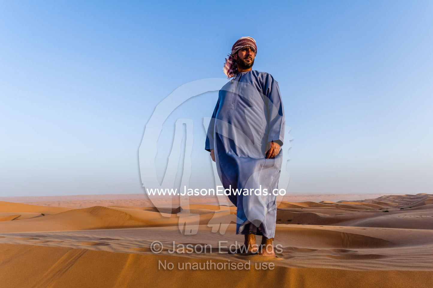 A Muslim man standing on a sand dune at sunset wearing a traditional dishdasha and masar. Wahiba Sands, Ramlat al-Wahiba, Sharqiya Sands, Sultanate of Oman.