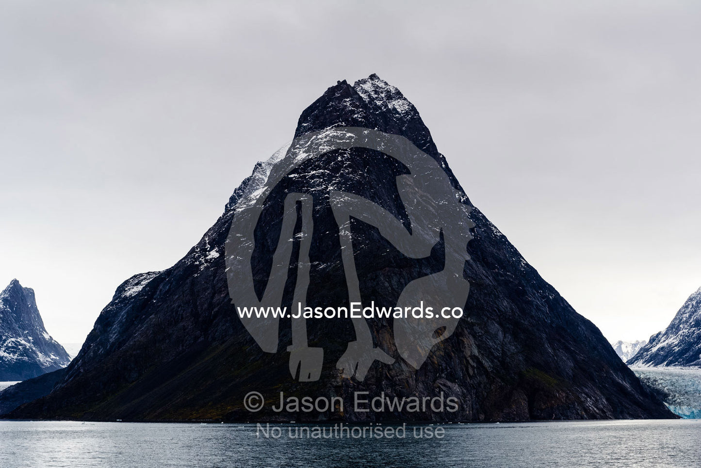 A cone-shaped mountain rises from the dark waters of an Arctic fjord beneath a storm laden sky. Fjord of Eternity, Maniitsoq, Sukkertoppen, Qeqqata Municipality, Greenland.