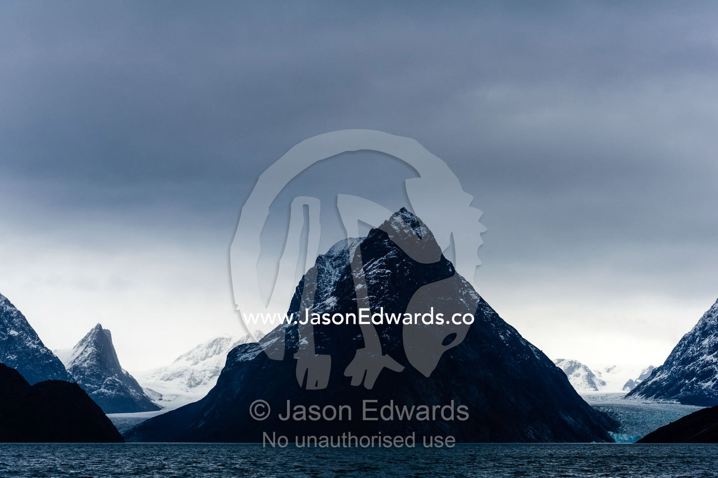 A cone-shaped mountain rises from the dark waters of an Arctic fjord beneath a storm laden sky. Fjord of Eternity, Maniitsoq, Sukkertoppen, Qeqqata Municipality, Greenland.