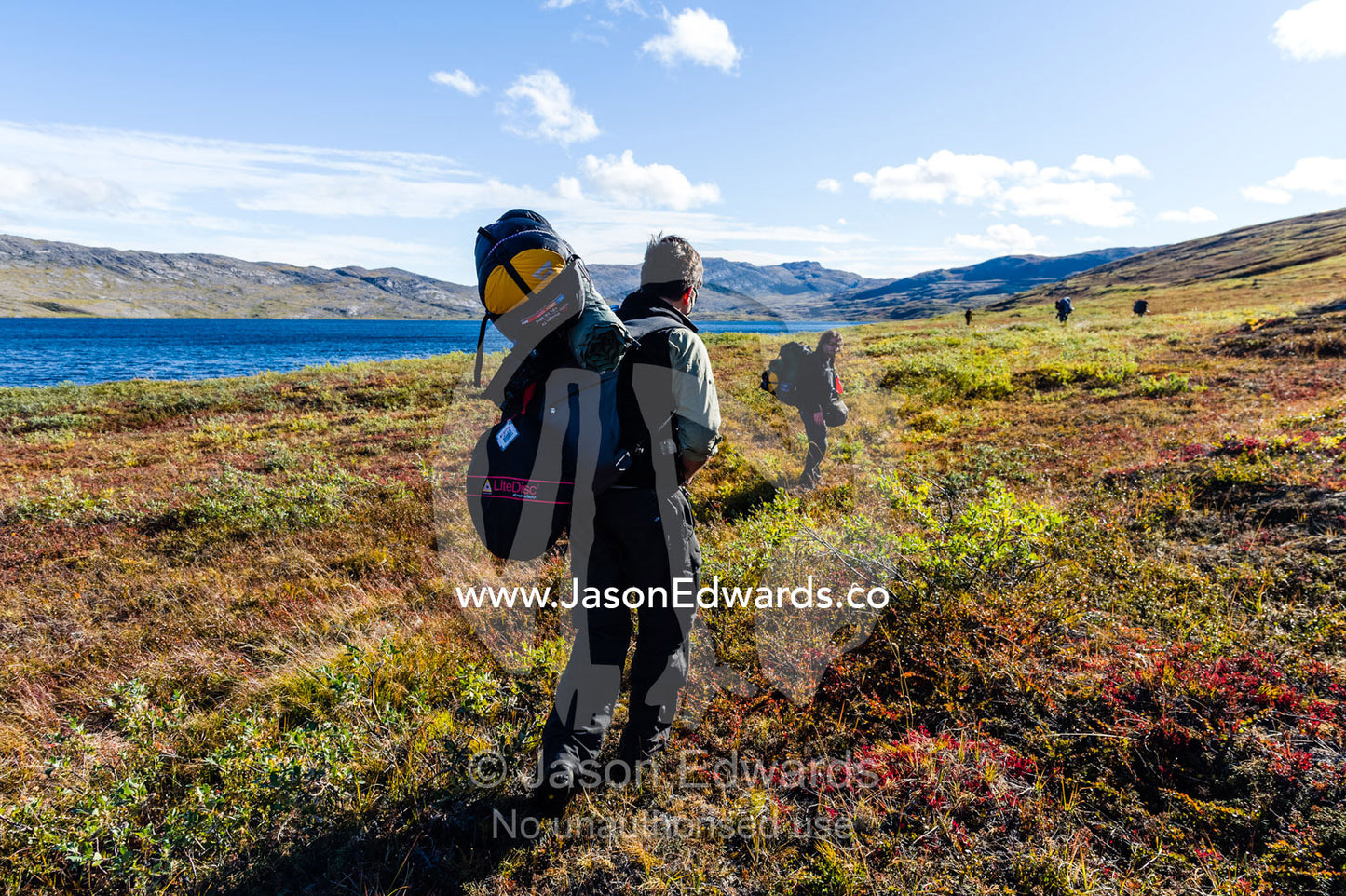 Hikers carrying backpacks traverse the tundra along shoreline of an alpine lake. Tasersuatsiaq Lake, Kangerlussuaq, Qeqqata Municipality, Greenland.