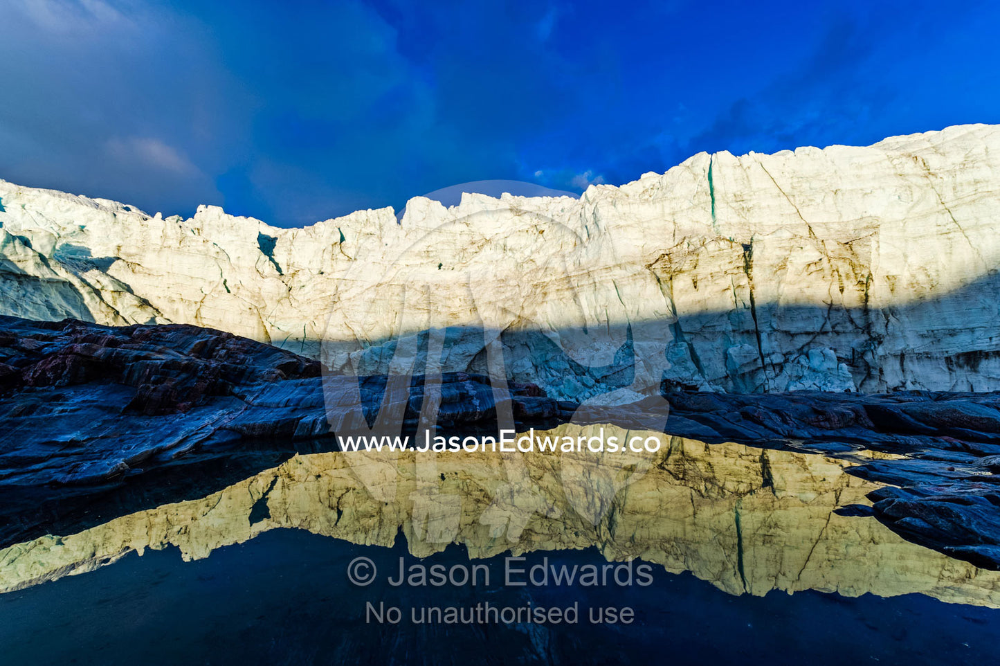 The setting sun reflects the sheer ice cliff of a glacier fracture zone in a pond. Russell Glacier, Greenland Ice Sheet, Qeqqata Municipality, Kangerlussuaq, Greenland.