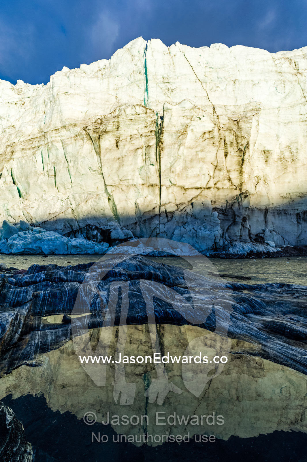 The setting sun reflects the sheer ice cliff of a glacier fracture zone in a pond. Russell Glacier, Greenland Ice Sheet, Qeqqata Municipality, Kangerlussuaq, Greenland.