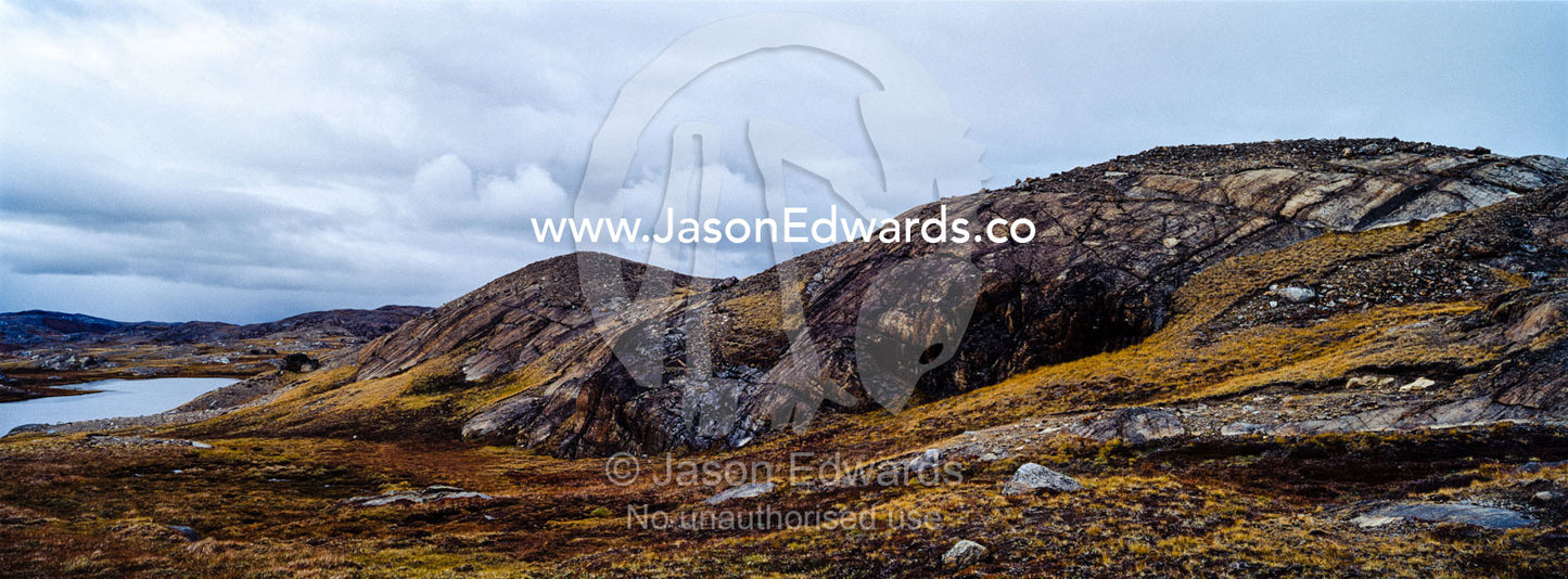Outcrops of exposed barren rocks emerge through the tundra surrounding an alpine lake. Isunngua Highland, Kangerlussuaq, Qeqqata Municipality, Greenland.
