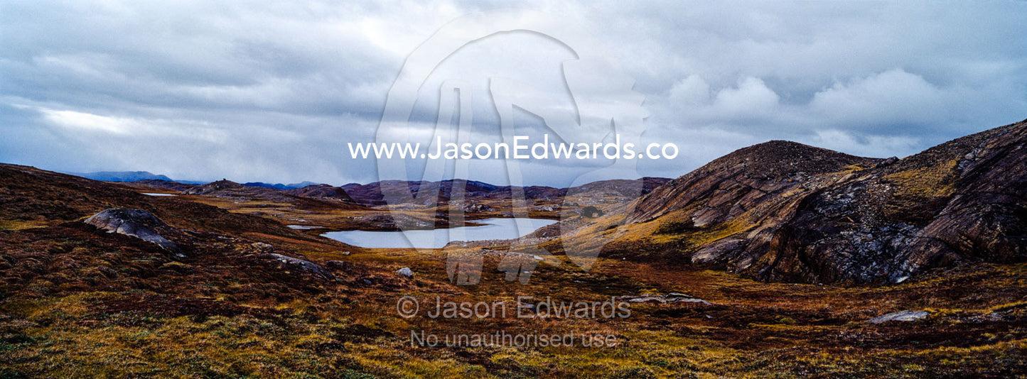Outcrops of exposed barren rocks emerge through the tundra surrounding an alpine lake. Isunngua Highland, Kangerlussuaq, Qeqqata Municipality, Greenland.