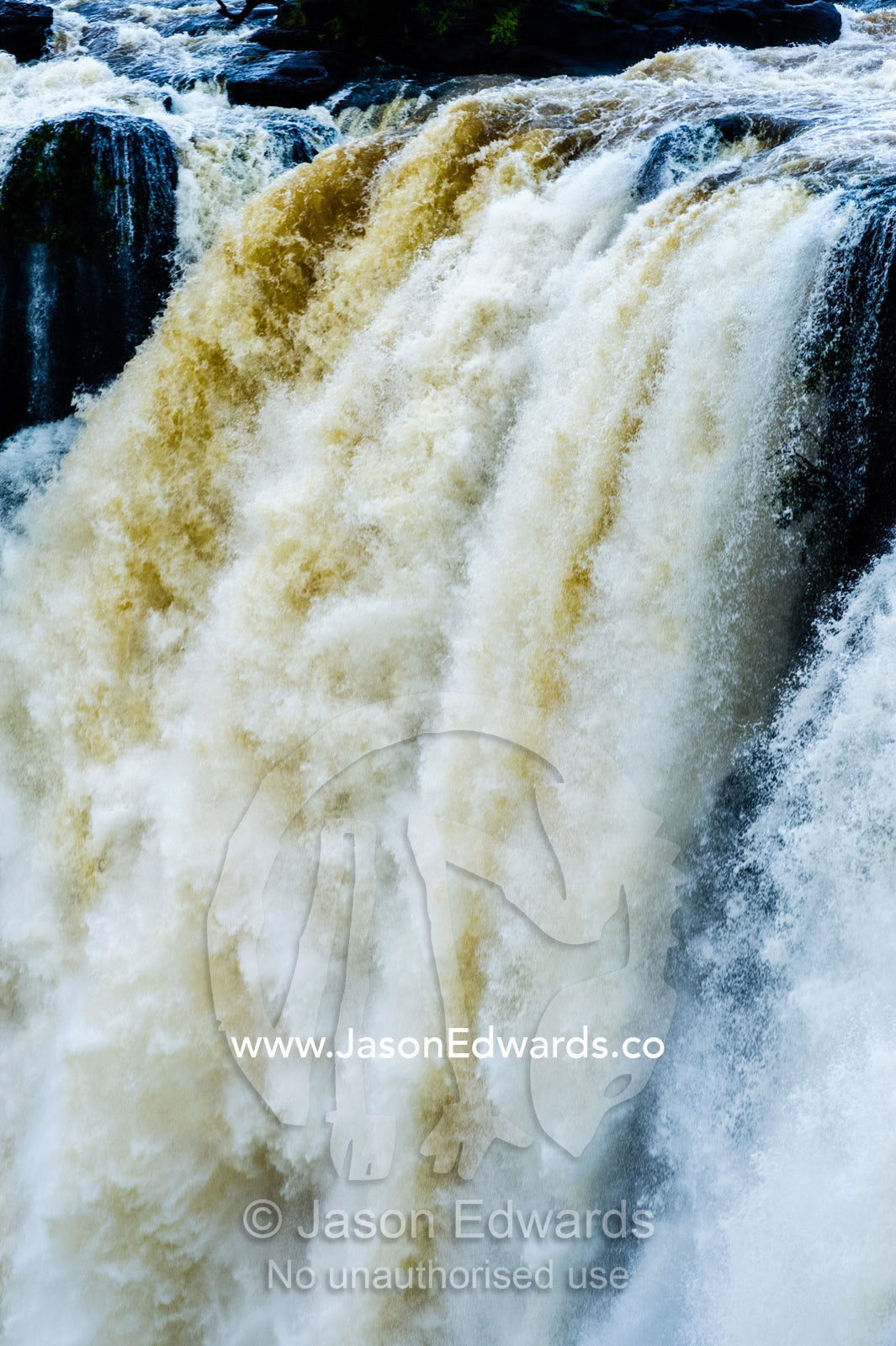 A flooded river cascades in a torrent over rocks on a waterfall lip. Zambezi River, Victoria Falls, Mosi-oa-Tunya National Park, Zambia.