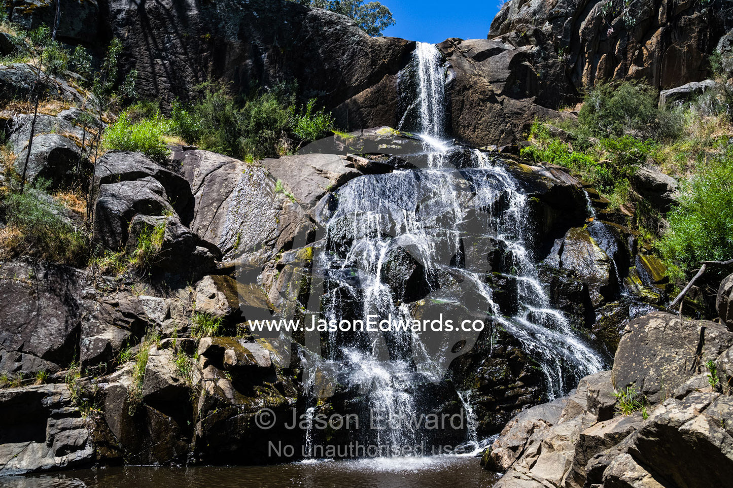 A stream cascades in a waterfall over boulders into a forest pool. Faithfuls Creek Waterfall, Euroa, Strathbogie, Victoria, Australia.