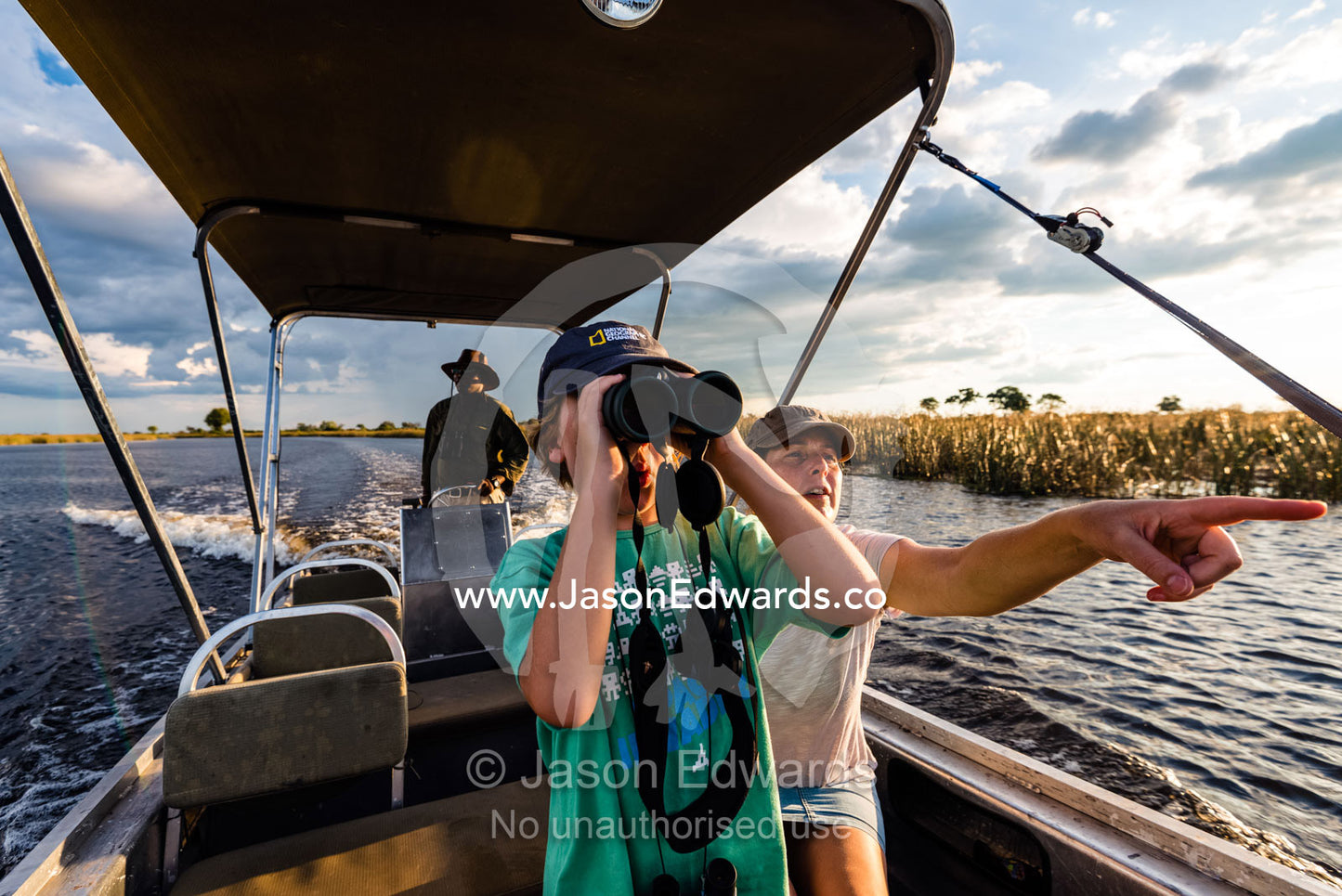 A mother and son watching river wildlife from a safari boat at sunset. Selinda Spillway, Selinda Reserve, Linyanti Region, Northern Botswana.