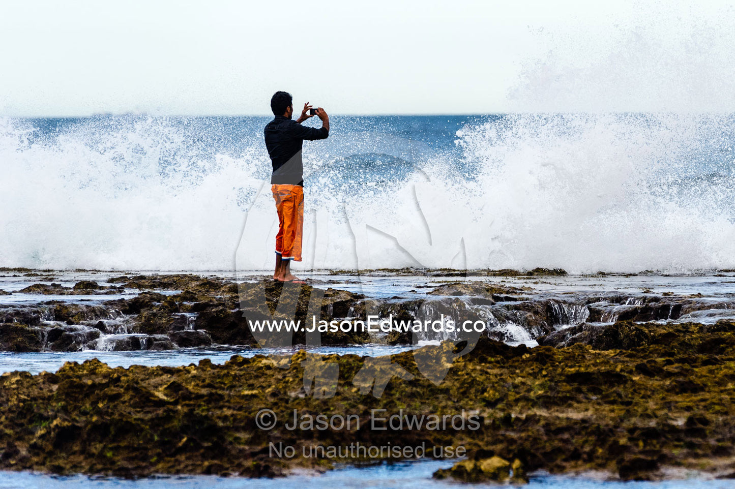 A man on a rock ledge takes a photo of crashing waves with a cell phone. Torquay, Surf Coast, Victoria, Australia.