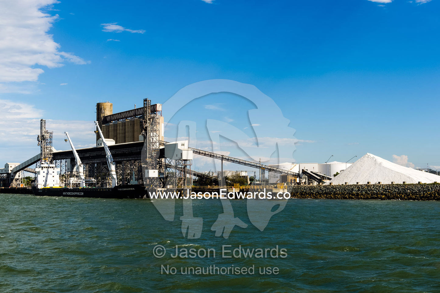 A project type ship loading wheat or bulk cargo. The stockpile of white material is calcite, used in industrial manufacturing, including toothpaste. Gladstone, Queensland, Australia.