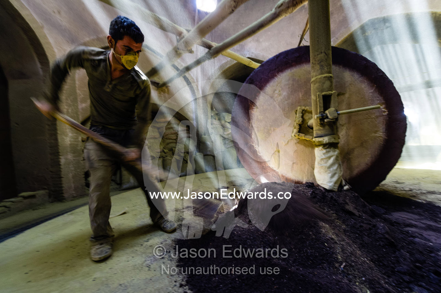 A labourer shovels henna seeds in front of a stone grinding wheel in a henna mill. Yazd, Yazd Province, Islamic Republic of Iran.