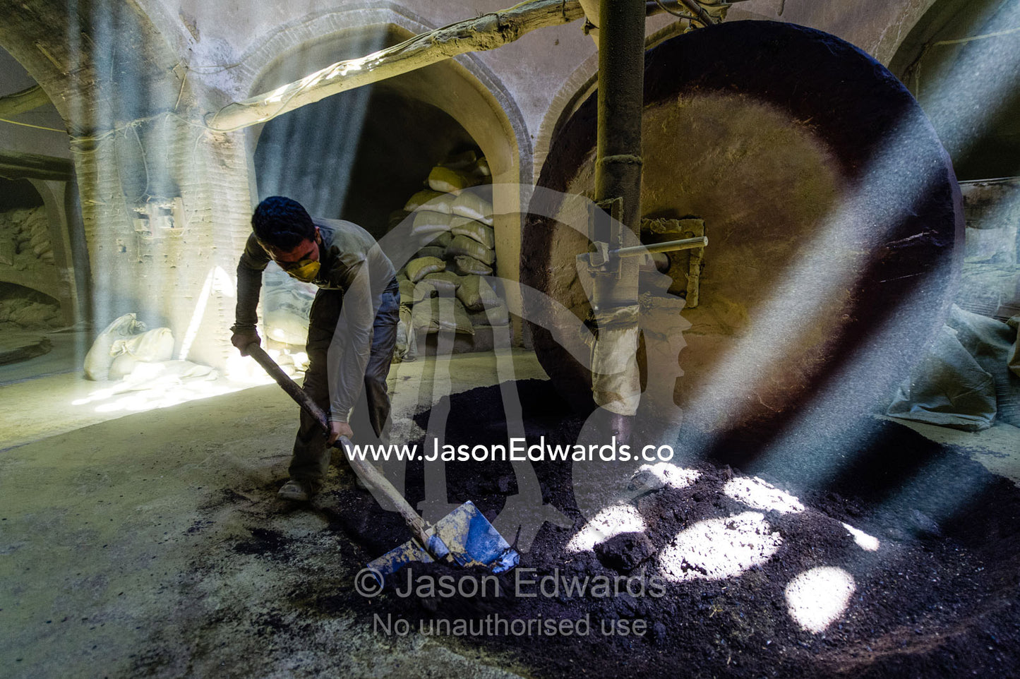 A labourer shovels henna seeds in front of a stone grinding wheel in a henna mill. Yazd, Yazd Province, Islamic Republic of Iran.