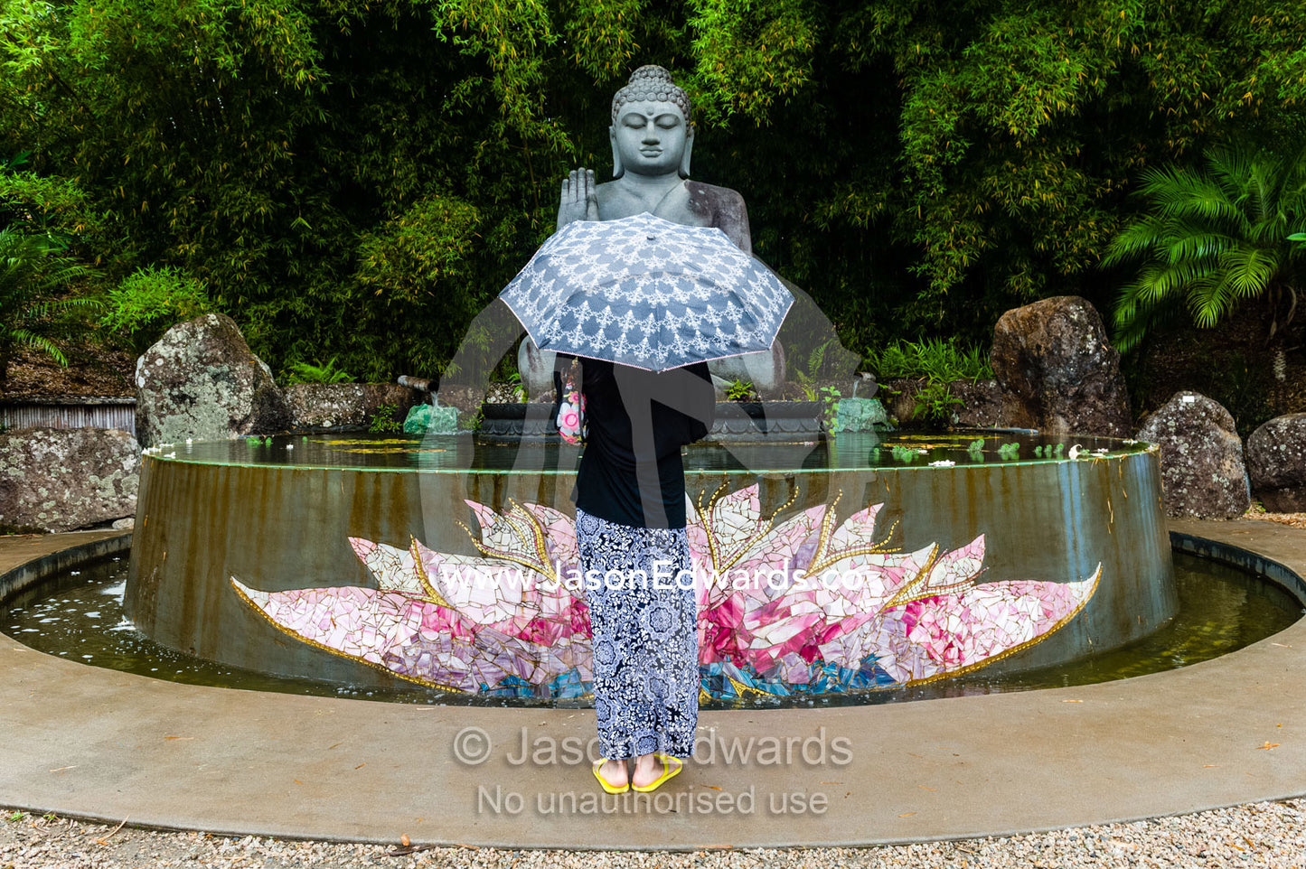 A tourist visits a statue of buddha offering a blessing over a lotus pond in a botanical garden. Crystal Castle, Shambhala Gardens, Mullumbimby, New South Wales, Australia.