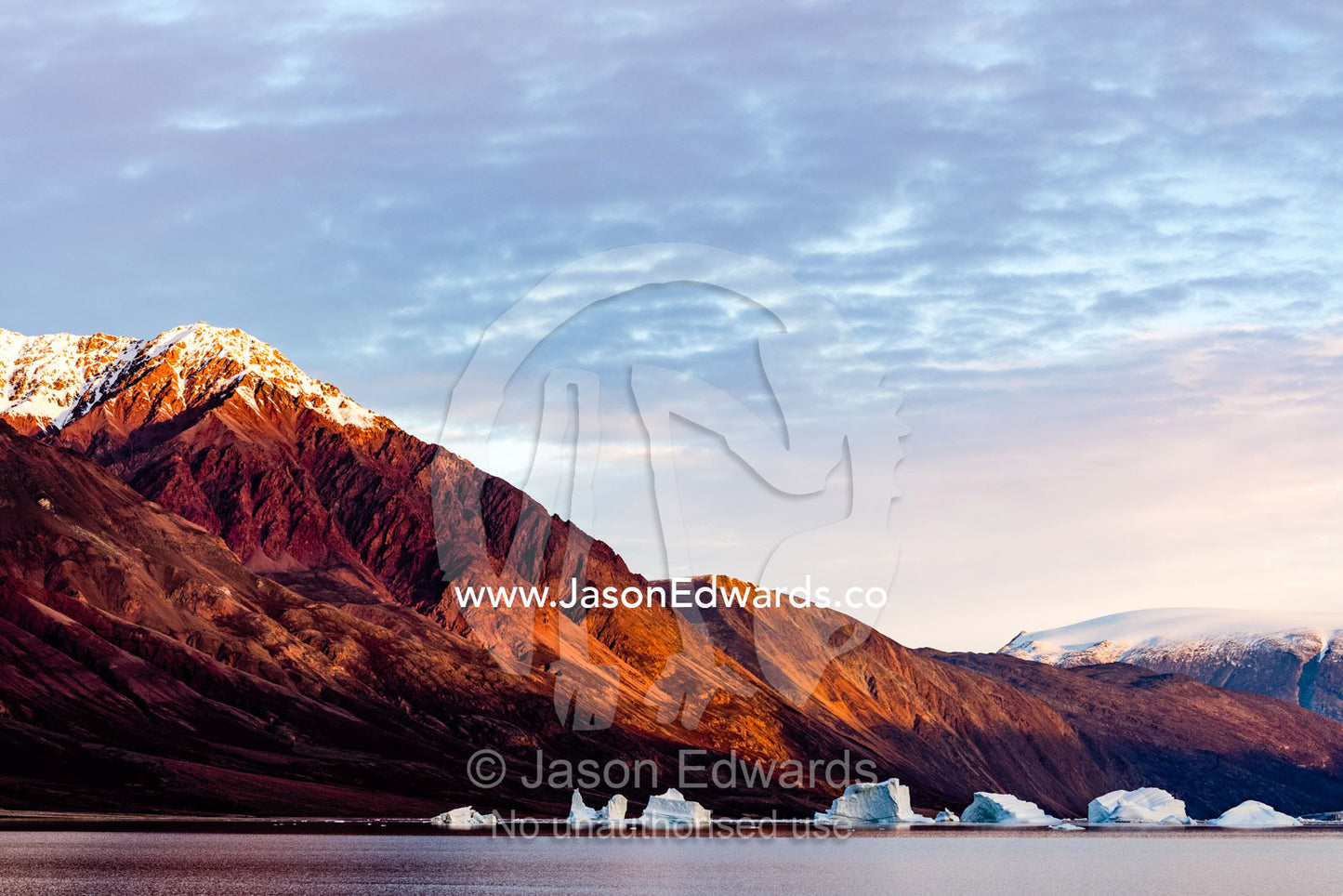 Dawn casts a blood-red glow over barren Arctic mountains surrounded by icebergs. Scoresby Sound, Greenland