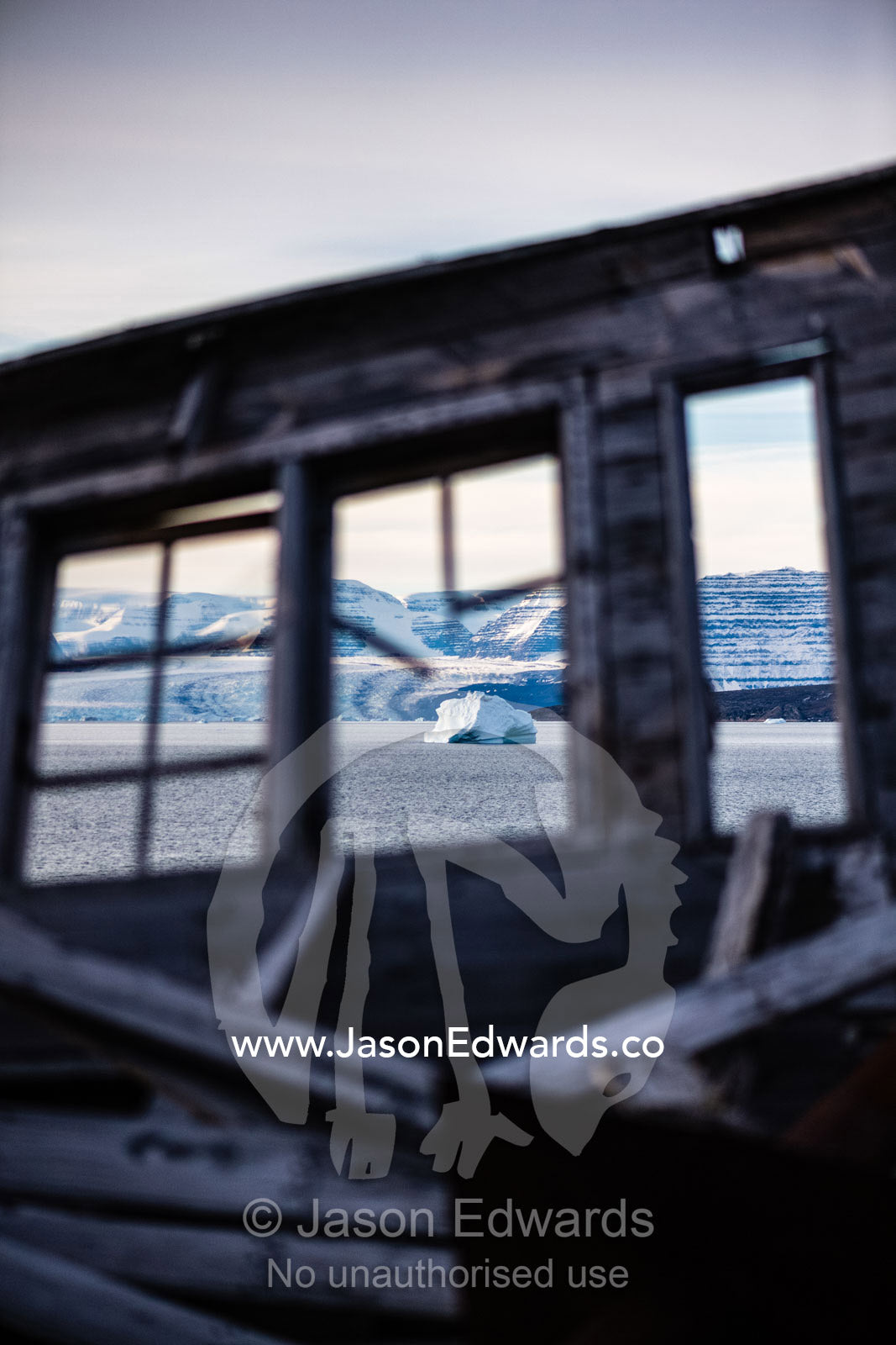 A weathered timber wall and window of a seal hunters hut looks over an iceberg in the arctic. Denmark Island, Hekla Havn, Scoresby Sound, Greenland