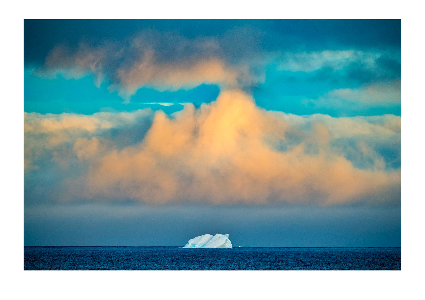 Pastel hues falling gently between tiered banks of laden storm clouds, caress a solitary iceberg in a vast ocean. Antarctic Peninsula, Antarctica.