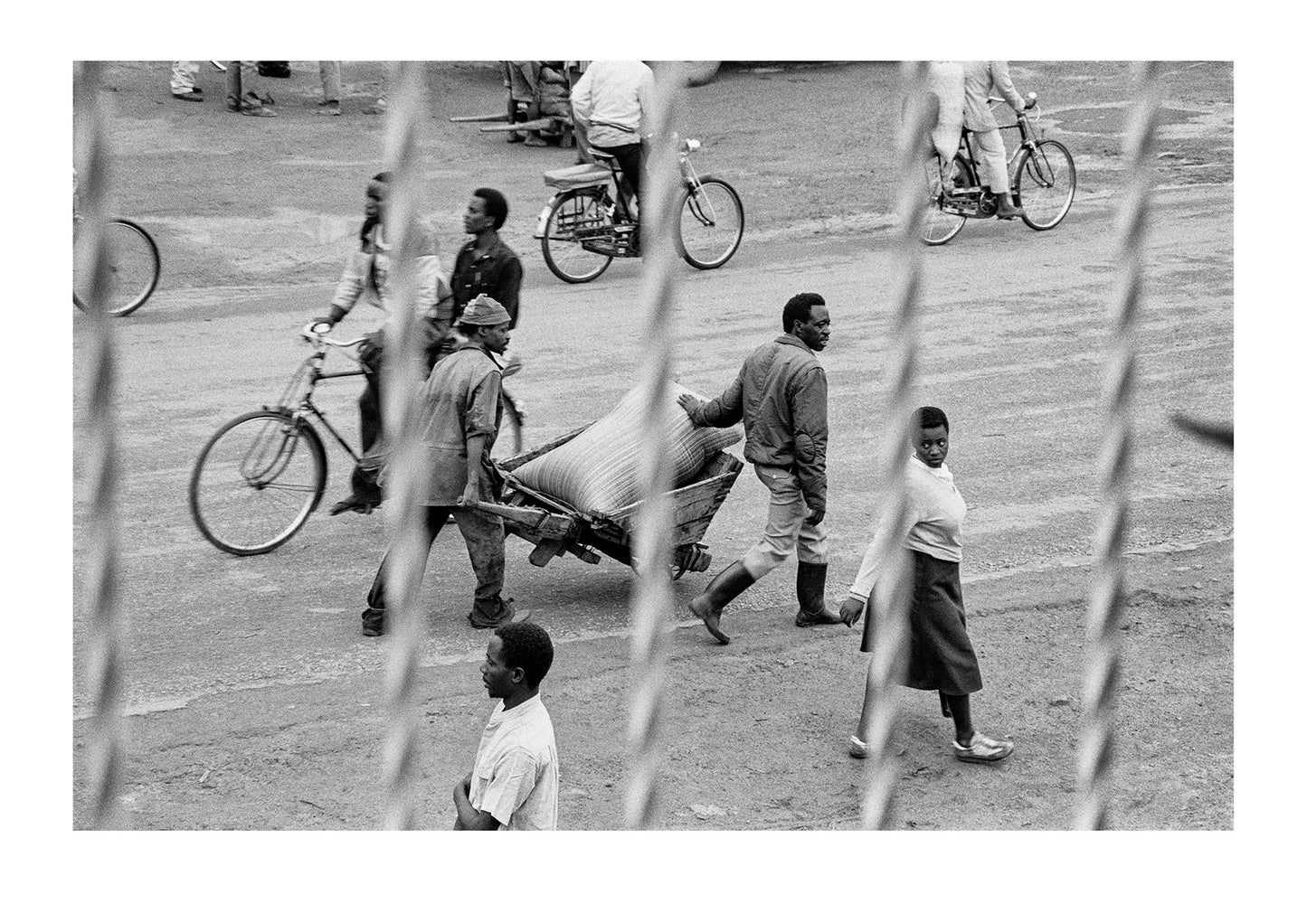 Labourers transport a bag of grain in a wheelbarrow whilst a lady casts a disapproving eye toward a passerby. Captured on Ilford HP5 black and white negative film. Kabale, Western Uganda.