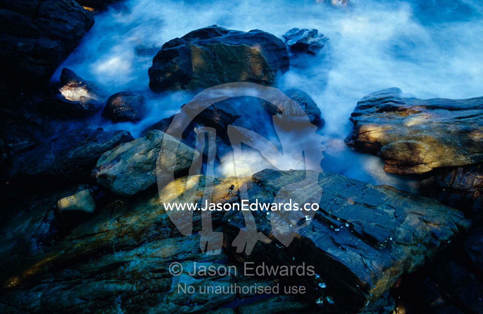 Churned up water cascades over rocks onto  a rocky, rugged shore. Bittangabee Bay, Ben Boyd National Park, New South Wales, Australia.