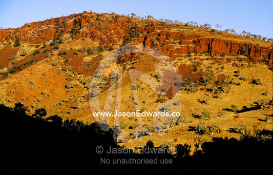 Sunset falls over a rich red volcanic cliff face and desert gorge. Hamersley Ranges Karijini National Park Western Australia