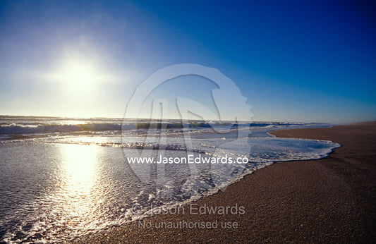 Gentle waves lap onto a pristine sandy beach with the sun reflecting. Coorong National Park, Younghusband Penninsula, South Australia.