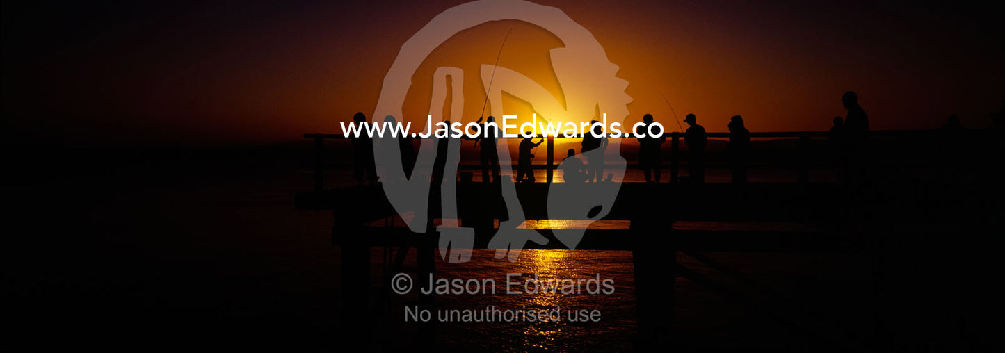 Holiday makers and fishermen in silhouette crowd a jetty at sunset. Merimbula, New South Wales, Australia.