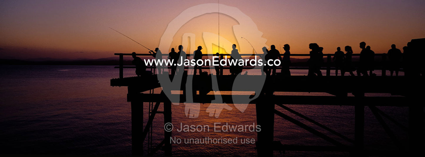 Holiday makers and fishermen in silhouette crowd a jetty at sunset. Merimbula, New South Wales, Australia.