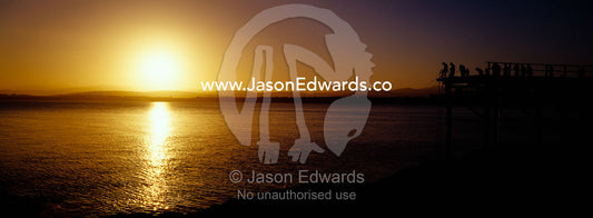 Holiday makers and fishermen in silhouette crowd a jetty at sunset. Merimbula, New South Wales, Australia.