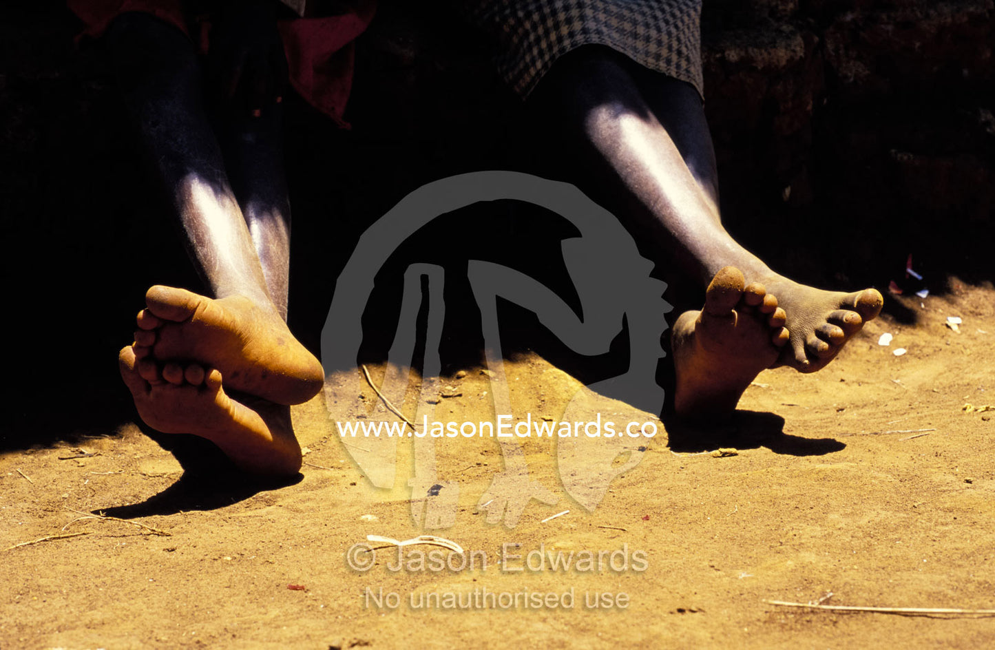 The feet of two teenage boys resting in the shade of a village hut. Tororo District, Uganda.