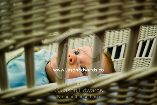 A small wide-eyed baby prepares for sleep in his wicker bassinet. East Malvern, Victoria, Australia.