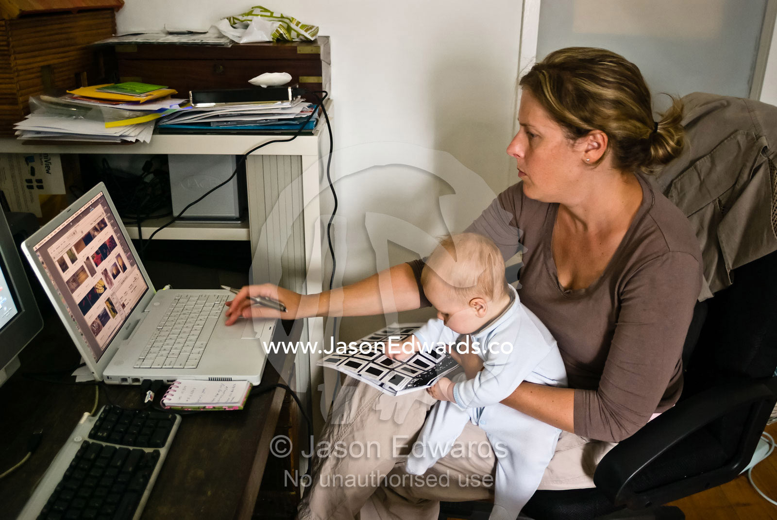 A new mother holds her young baby whilst working online in an office. Collingwood, Victoria, Australia.