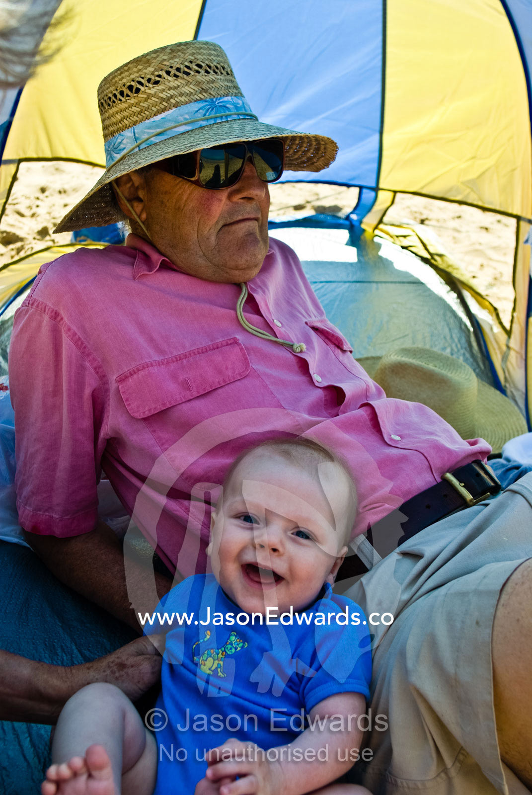A laughing baby shares a sun shade on the beach with his grandfather. Cosy Corner, Torquay, Victoria, Australia.