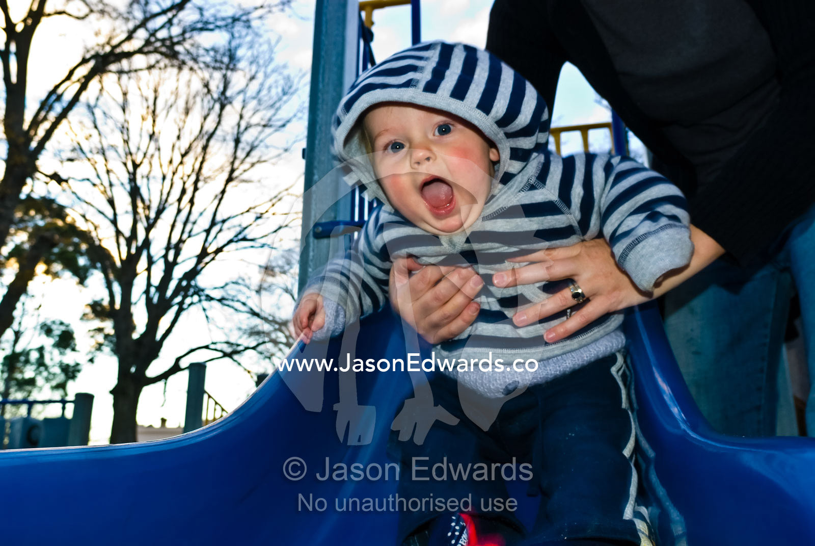 An excited baby shoots safely down a park slide in his mothers hands. Curtain Square, North Carlton, Victoria, Australia.