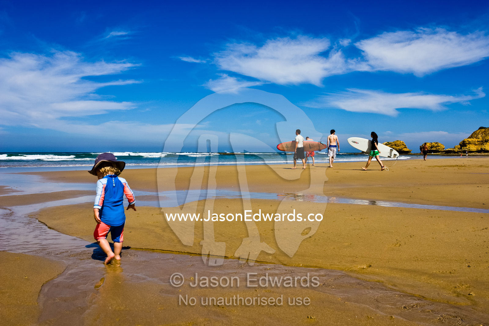 A baby explores a beach at low tide as surfers head for the surf. Torquay Point, Torquay, Surf Coast, Victoria, Australia.