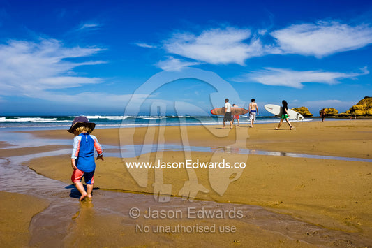 A baby explores a beach at low tide as surfers head for the surf. Torquay Point, Torquay, Surf Coast, Victoria, Australia.