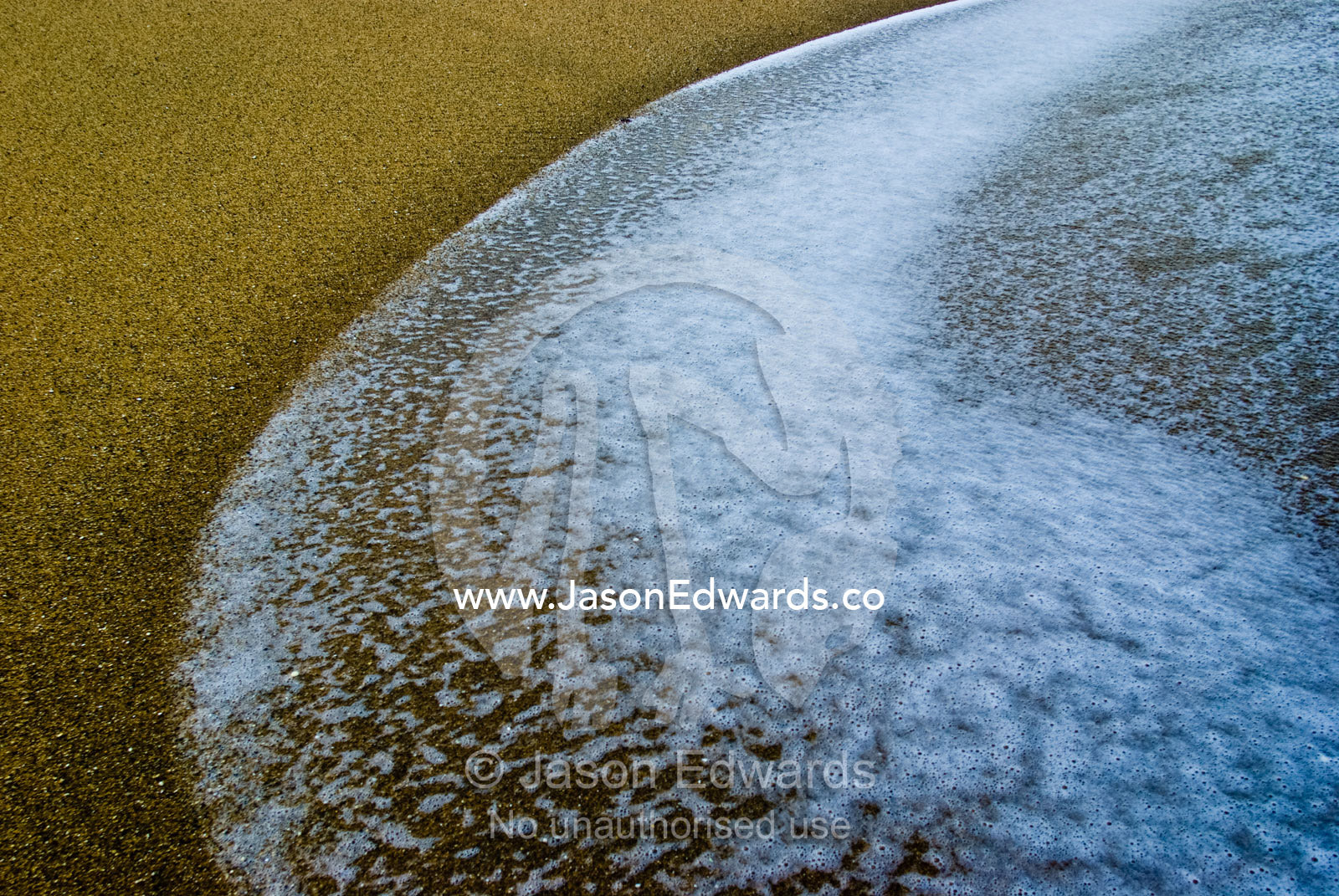 The foamy edge of a winter wave sweeps onto an empty sandy beach. Bells Beach Surfing Recreation Reserve, Surf Coast, Victoria, Australia.