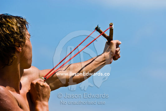 A young man fires a stone from a slingshot into the tropical blue sky. Bingin, Bali, Indonesia.