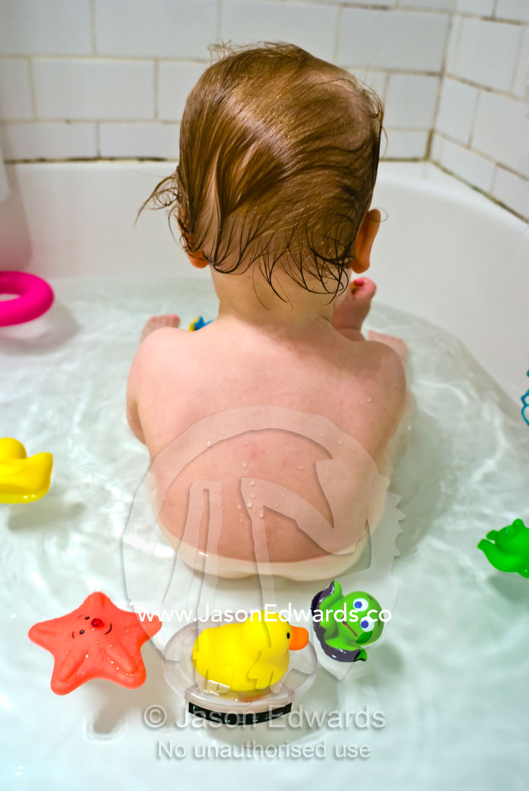 A fair-skinned baby sitting in a bath surrounded by his toys. North Carlton, Victoria, Australia.