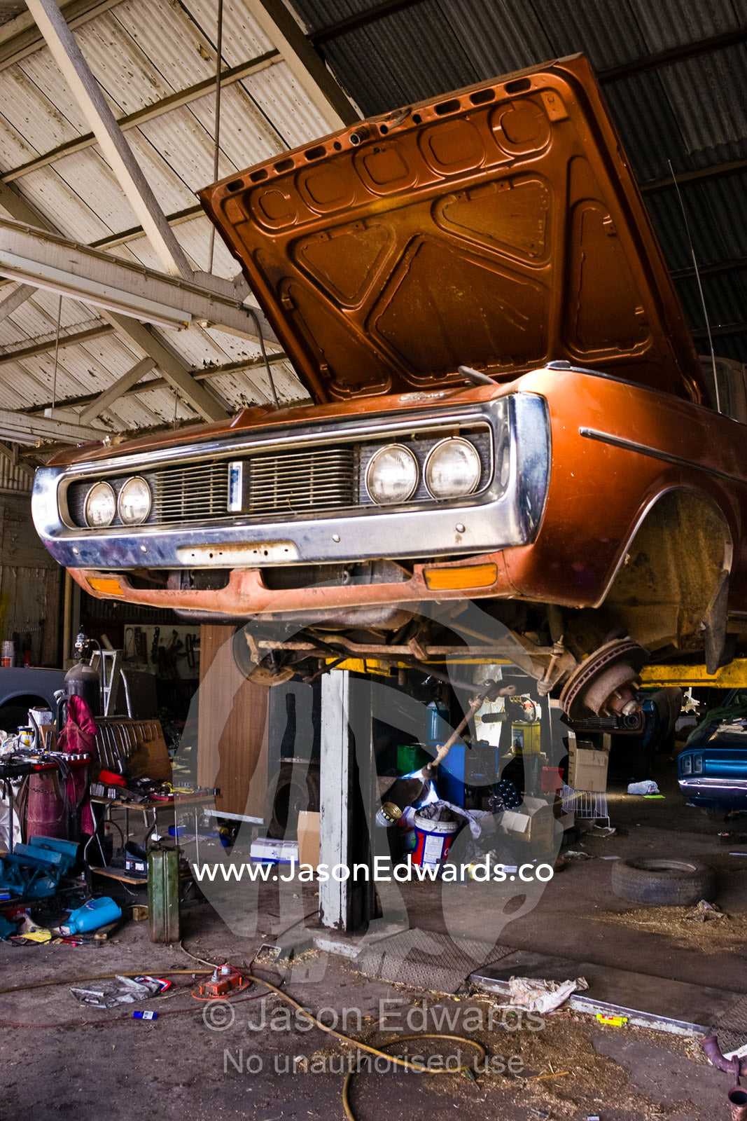 An old car rests on a hoist for repairs in a messy country workshop. Inglewood, Victoria, Australia.