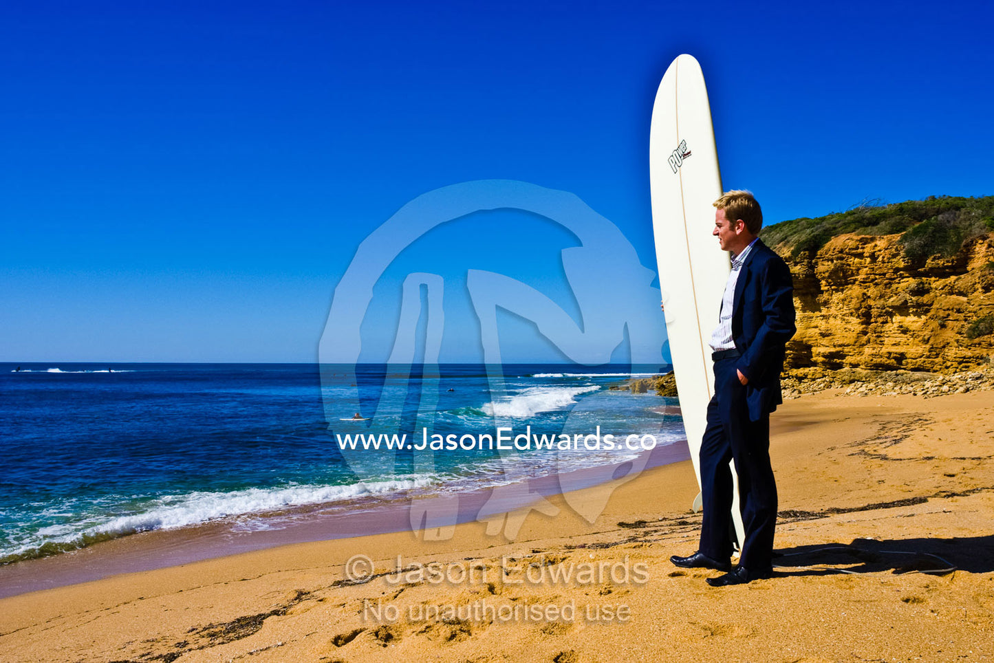 A businessman in a suit waits on the beach to go longboard surfing. Bells Beach Surfing Recreation Reserve, Victoria, Australia.