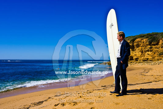 A businessman in a suit waits on the beach to go longboard surfing. Bells Beach Surfing Recreation Reserve, Victoria, Australia.