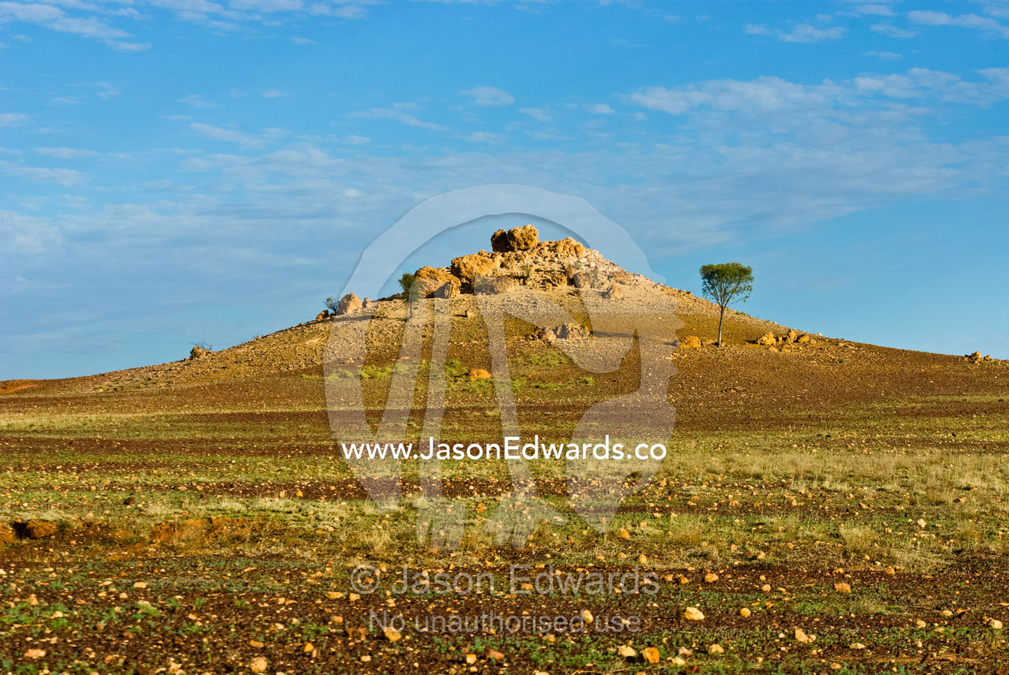 A boulder-strewn hill rises from a rugged desert plain at sunset. Cramsie Muttaburra Road, Queensland, Australia.