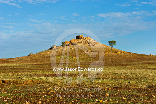 A boulder-strewn hill rises from a rugged desert plain at sunset. Cramsie Muttaburra Road, Queensland, Australia.