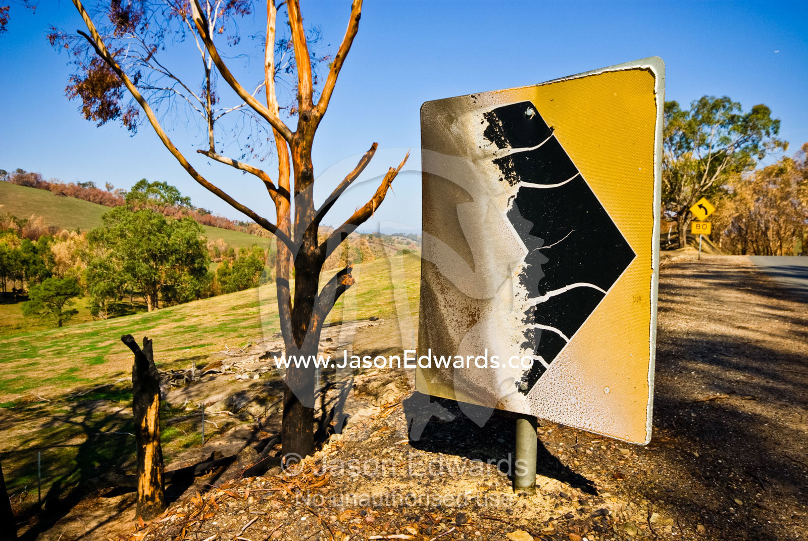 A fire damaged metal safety sign on a bend in a country road. Junction Ridge Road, near Kinglake National Park, Victoria, Australia