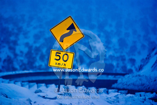 A speed and road warning sign on a steep snow-covered mountain pass. Great Alpine Road, Alpine National Park, Great Dividing Range, Victoria, Australia.