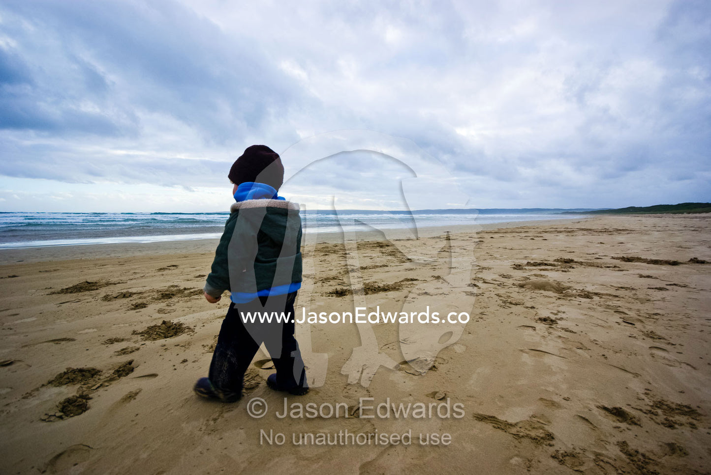 A small boy explores a cold, grey sweeping ocean beach in winter. Sandy Point, Victoria, Australia