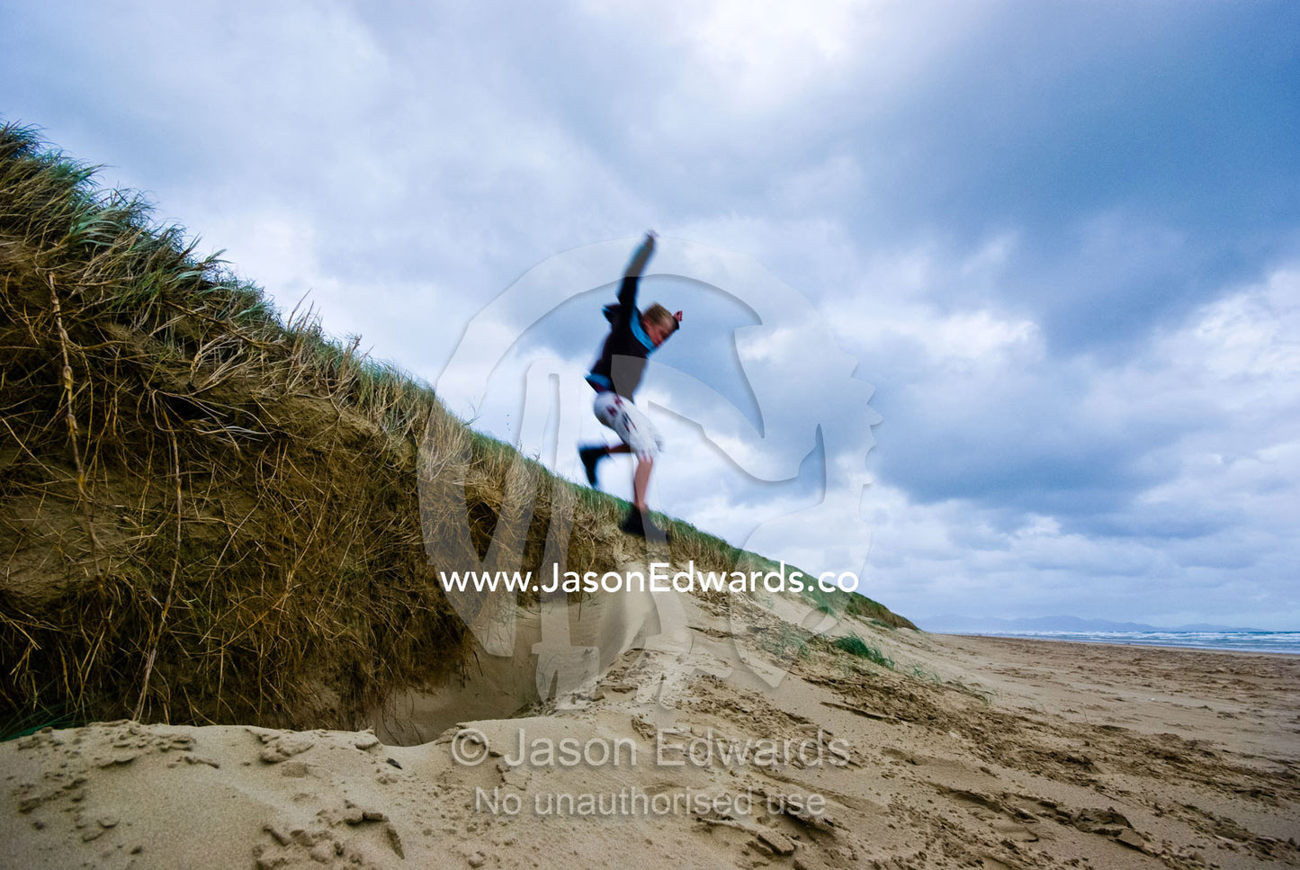 A boy leaps from a sand dune on a grey and stormy winters day. Sandy Point, Victoria, Australia