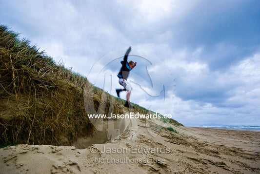 A boy leaps from a sand dune on a grey and stormy winters day. Sandy Point, Victoria, Australia