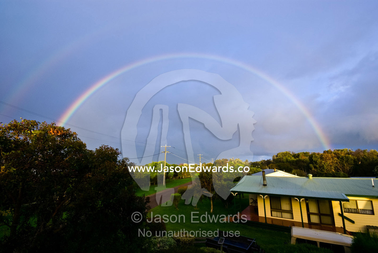 A rainbow arches over houses in a small tree-lined suburban street. Sandy Point, Victoria, Australia