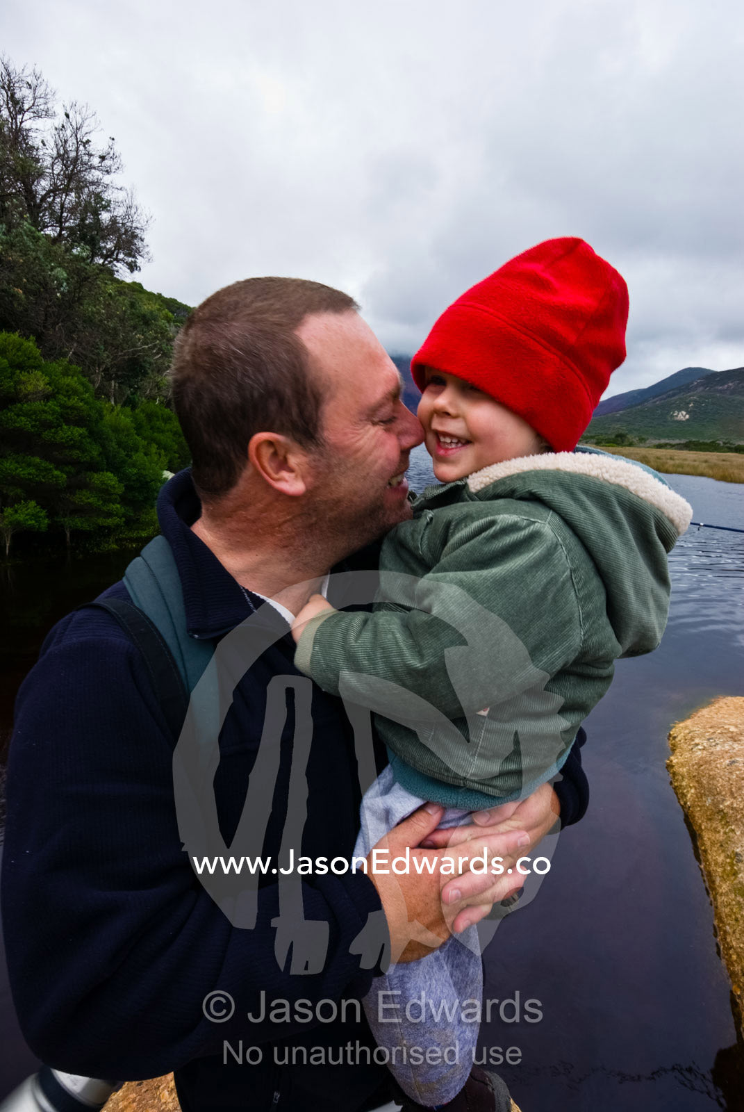 A father cuddles his small son by a river on a winters day. Tidal River, Wilsons Promontory National Park, Victoria, Australia
