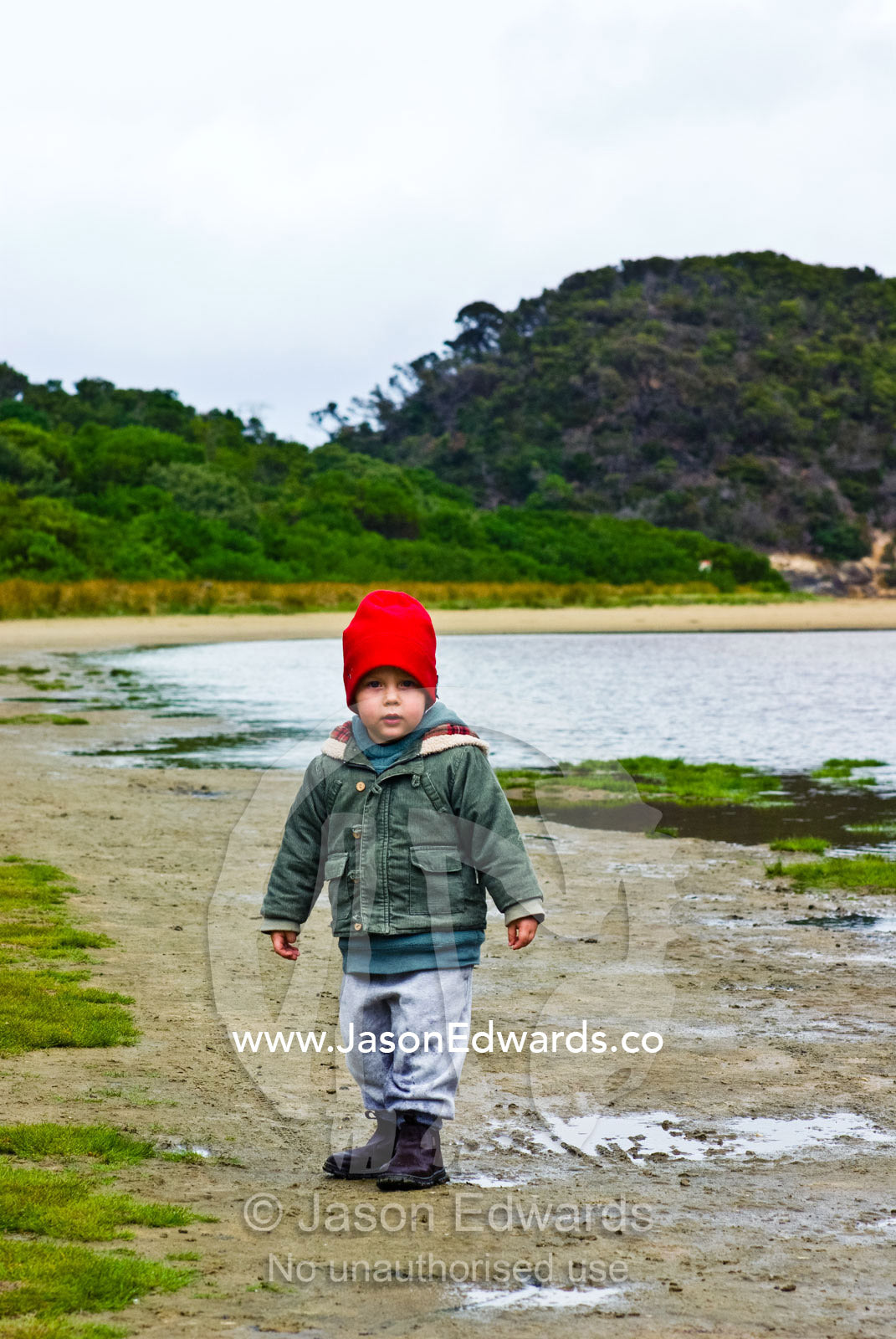 A small boy dressed in winter clothes walking along a river shore. Tidal River, Wilsons Promontory National Park, Victoria, Australia